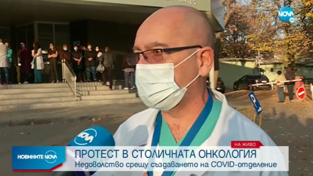 Директорът на онкологията Стефан Константинов дава интервю, а зад гърба му се виждат протестиращите лекари от болницата